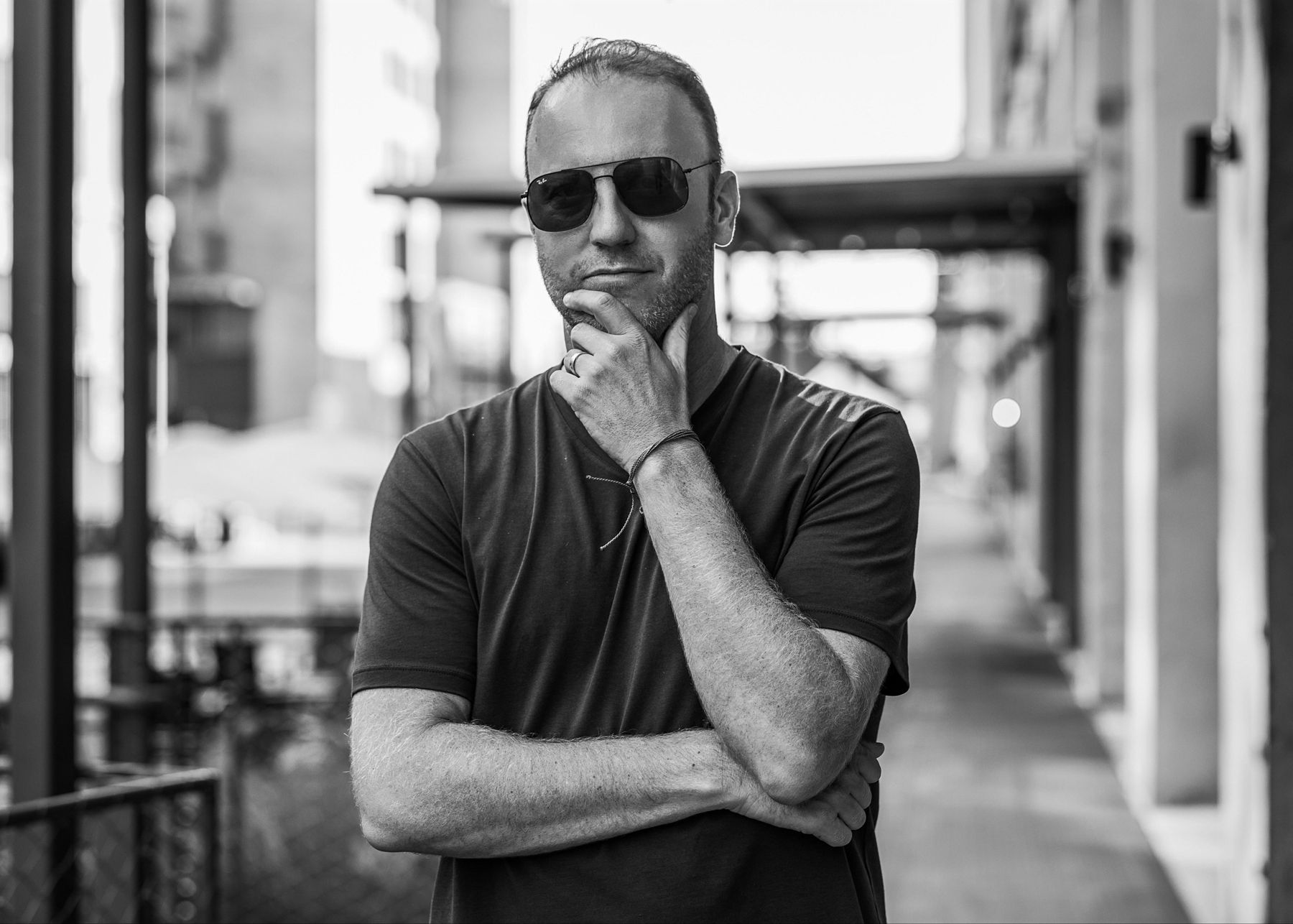 Darren Marble in sunglasses and a black t-shirt, hand on chin, on a city sidewalk